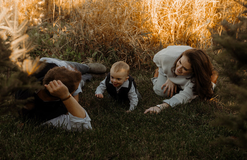 A family of three enjoys a sunny day amidst the tall, golden grasses, captured beautifully by a New England portrait photographer. The man, woman, and their crawling toddler share smiles and loving gazes in this picturesque outdoor setting.