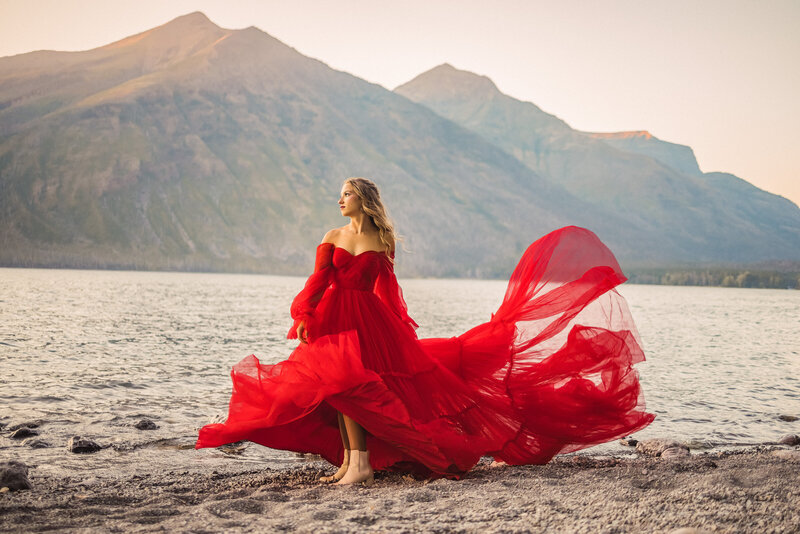 blonde senior girl in a long flowing red gown standing in the wind by the water with large mountains in the background