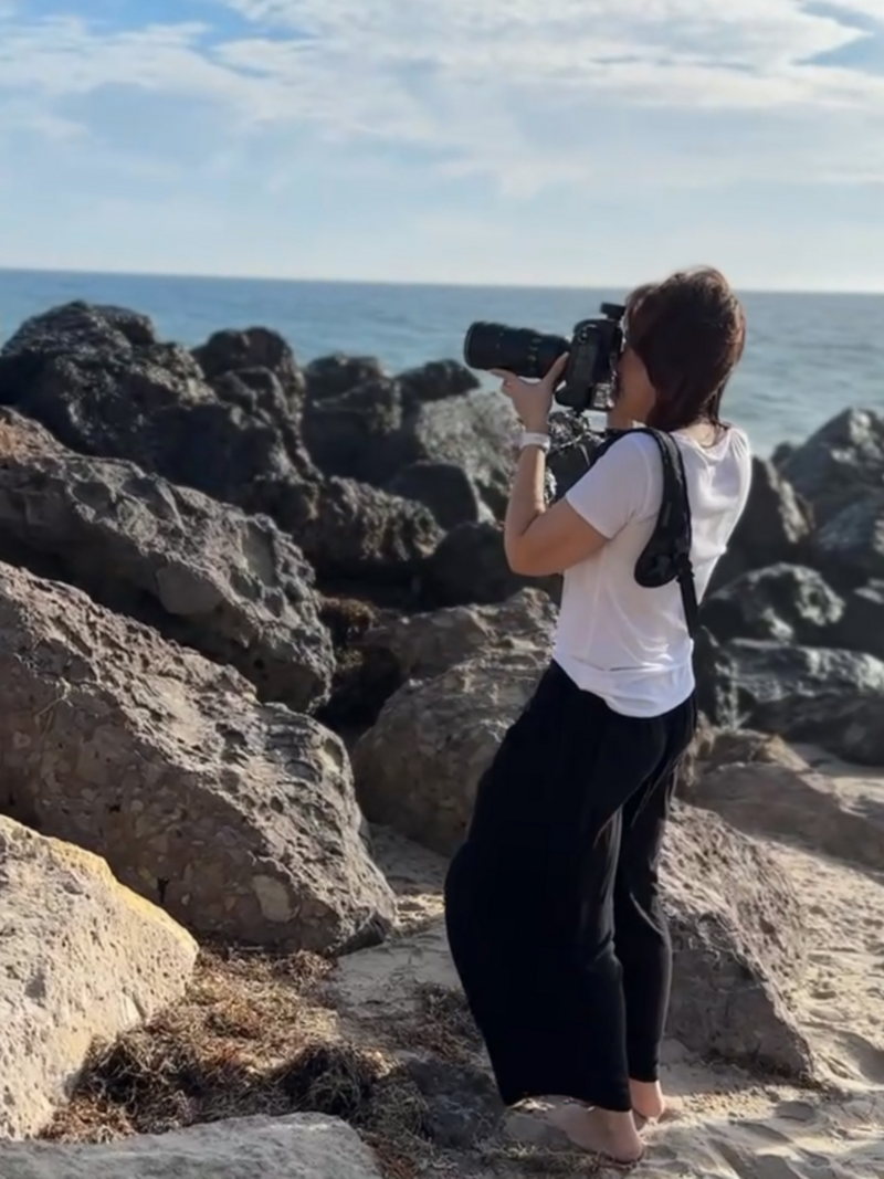 los angeles photographer shooting at the beach woman holding nikon camera on rocks with ocean in the background and blue sky with clouds