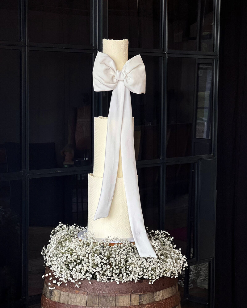 Three tier wedding cake with an illusion floating tier with large white bow  sitting on a cloud of white Gypsophilia set up at The Barn at Botley Hill Surrey