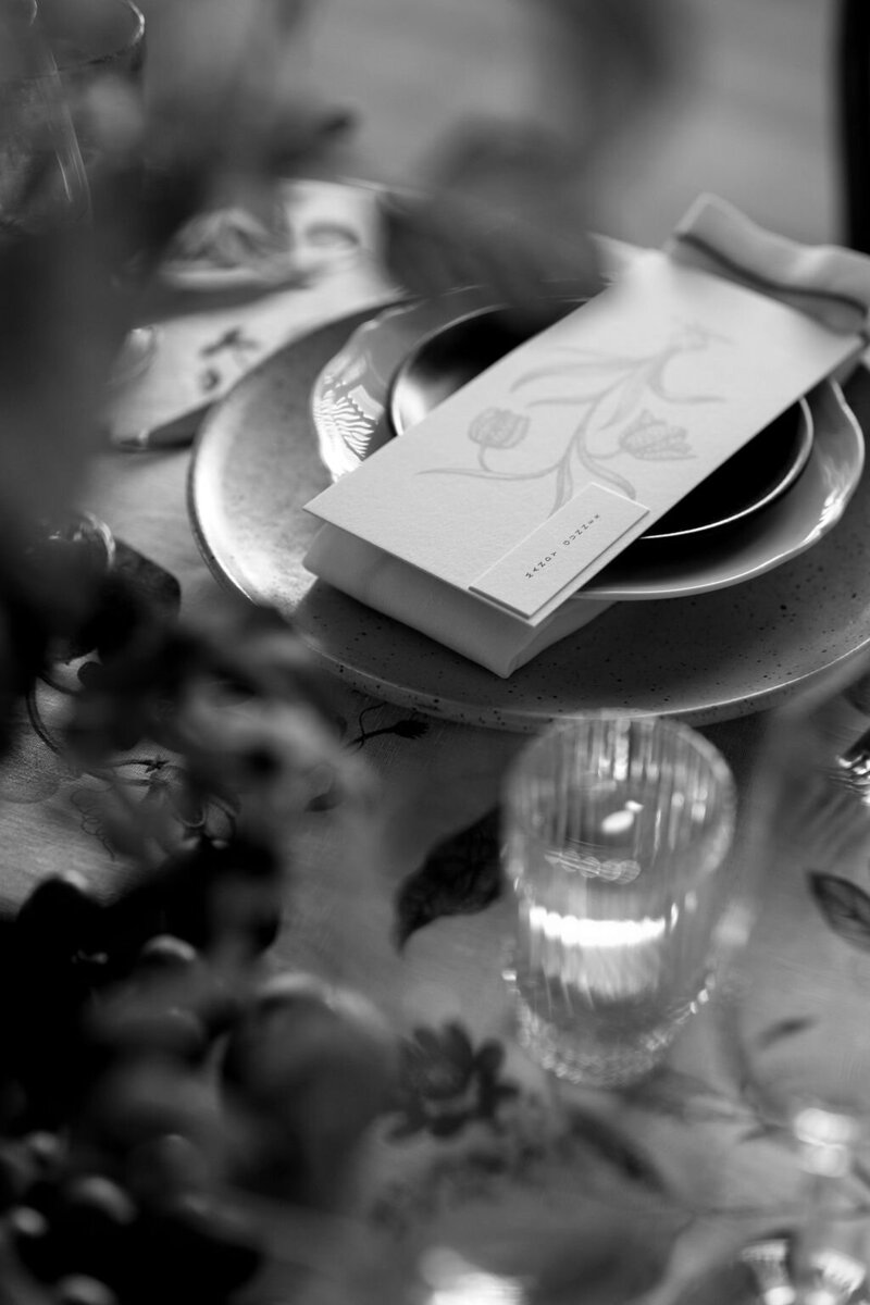 A black-and-white close-up of a place setting with a floral-illustrated menu, layered plates, and a glowing candle.