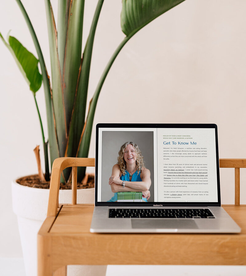 A website for Heidi Schauster on a laptop screen rests on a table next to a green plant, creating a cozy atmosphere.