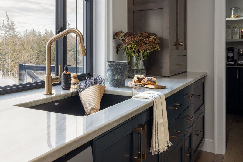Kitchen sink with gold faucet. Main cabinets are a deep blue and upper cabinets are dark oak. There is a bouquet of lavender in the sink and chocolate covered croissants on the counter.