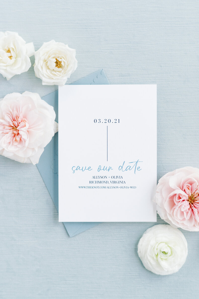 Dusty blue save the date card displayed on a blue styling mat with flowers.
