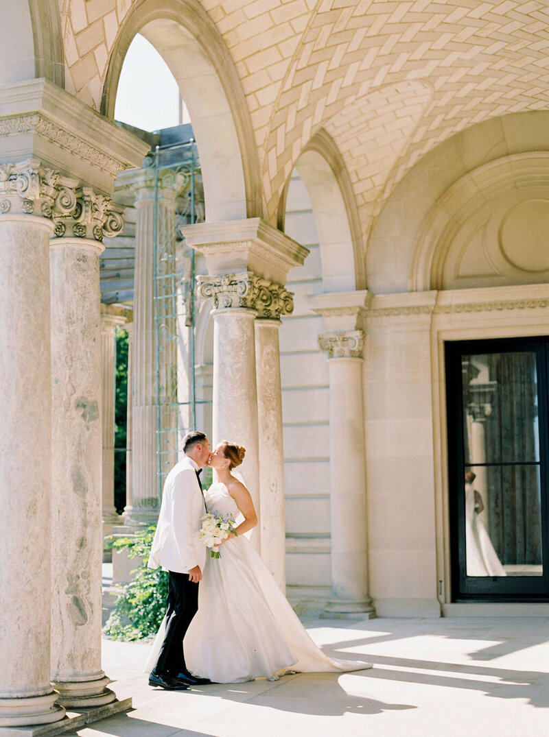 Wedding Photographer RI | A bride and groom share a kiss under an ornate stone archway. She wears a white gown, holding a bouquet, while he is in a white suit.