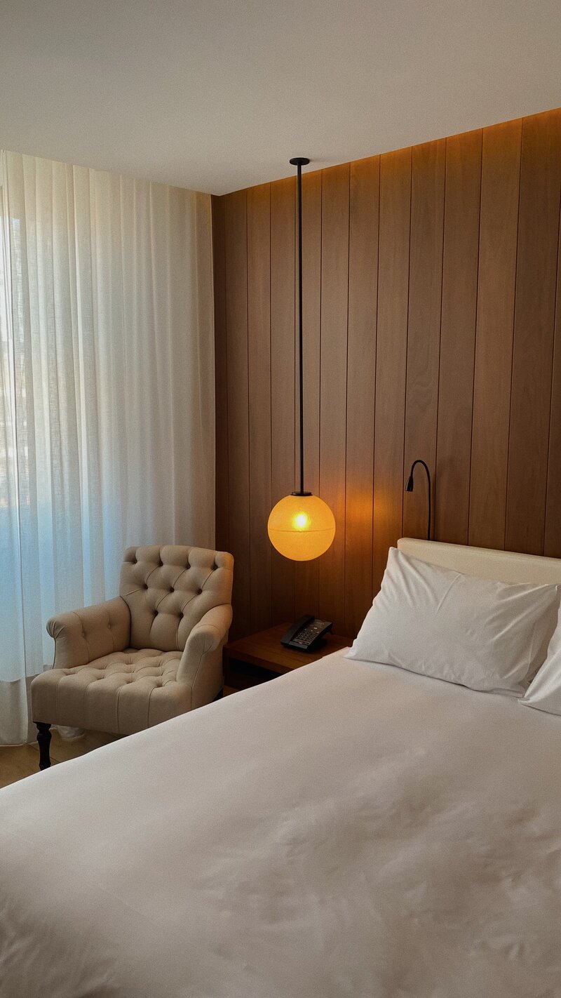 The interior of a hotel room. Pictured is the bed, light fixture, and chair