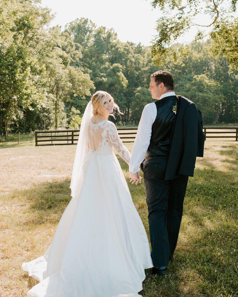 bride and groom holding hands walking in a green pasture with horses in Richmond, Virginia