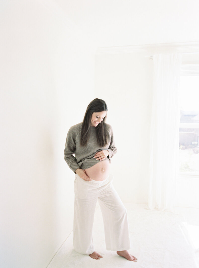 Brunette, pregnant mother sweater and white pants shows a peak of her baby belly taken by photographer Little Rock Bailey Feeler.
