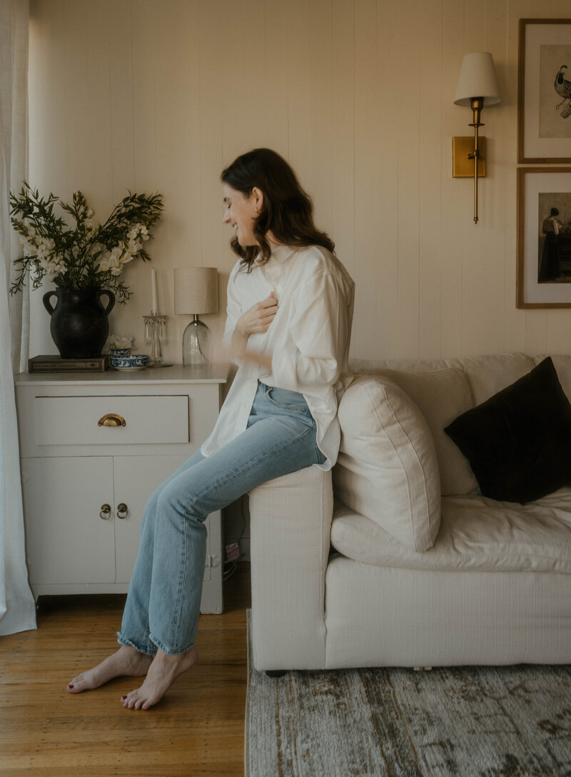 Woman wearing white button down and blue jeans sitting on the arm of a cream couch and looking to the side