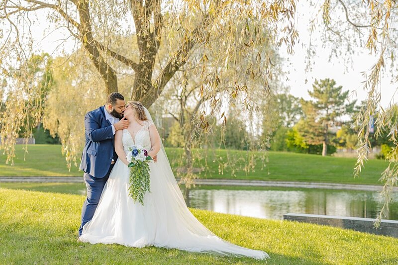 Bride and groom kissing under willow tree by pond at SVSU