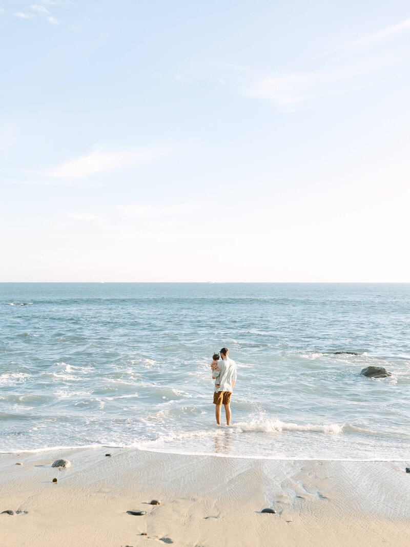 A dad stands at the edge of the ocean holding a child. The sky is clear and the waves gently lap at their feet, conveying a serene and peaceful mood.
