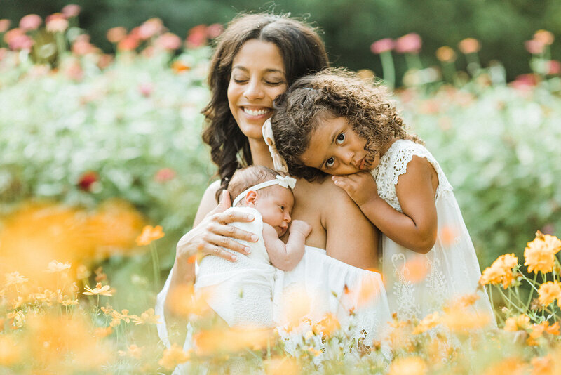 mom holds baby and is hugged by young daughter in vibrant flower garden