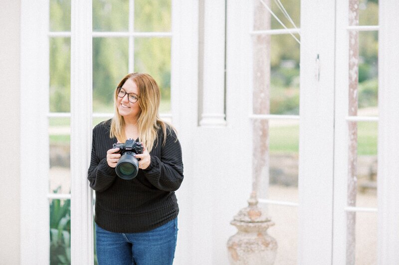 Colour image of Lisa Peel Photography , standing in Came House, Dorset, holding her camera. Wearing a black jumper and jeans