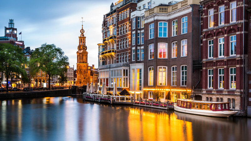 A calm canal reflecting warm lights from tall historic buildings at dusk, with a clock tower in the distance and trees along the waterfront.