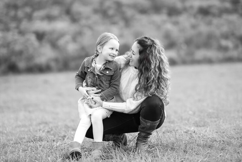 Mom is crouching down with little girl sitting on her knee. Mom and daughter are smiling at each other in Philadelphia, PA park.