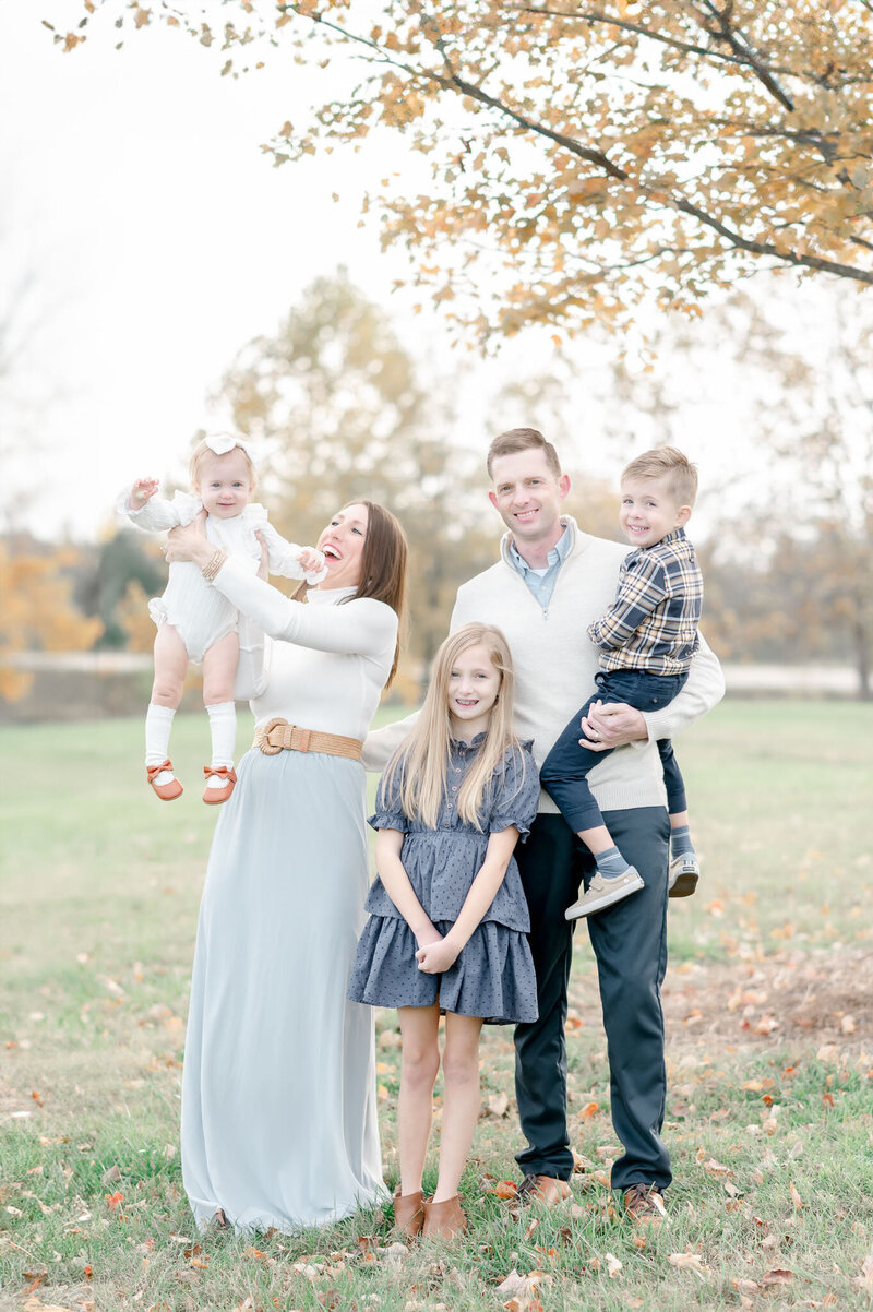 A mom holds her baby in the air while the rest of the family smiles