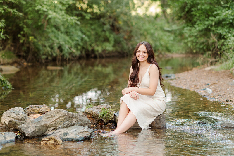 Senior girl wearing a cream dress sitting on stones in a creek by Lancaster PA photographer