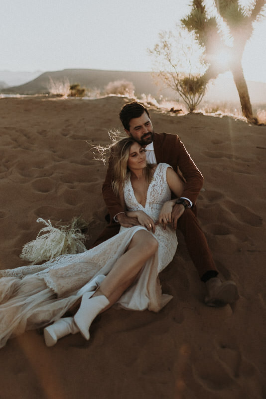 A boho elopement couple with a groom in a rust suit and a bride in a lace wedding dress sitting on the ground at sunset in Joshua Tree.