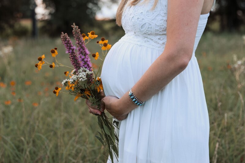 Woman in white dress holding her pregnant belly with a bouquet of wildflowers