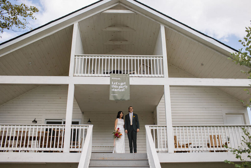 Bride and groom stand at top of gray steps under a white A-frame building with a banner that says "let's get this party started!"