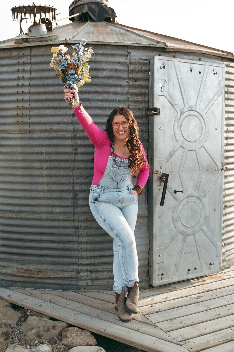 Stacey Domolewski, a rural brand photographer stands near a grainbin with flowers 