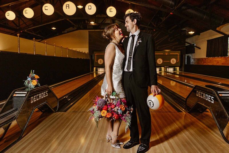 A bride and groom celebrate at Decades vintage arcade in Lancaster on their elopement day. They are posing in the bowling alley, and the bride is holding her bouquet and the groom is holding a bowling ball