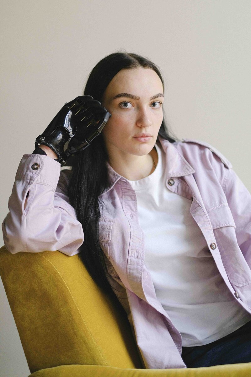 A young person with long, black hair looks into the camera with a neutral expression. They are wearing a long-sleeve, purple denim shirt and are sitting in a yellow velvet chair. Their right arm is propped up on the top of the chair, and their black prosthetic hand rests against their cheek.