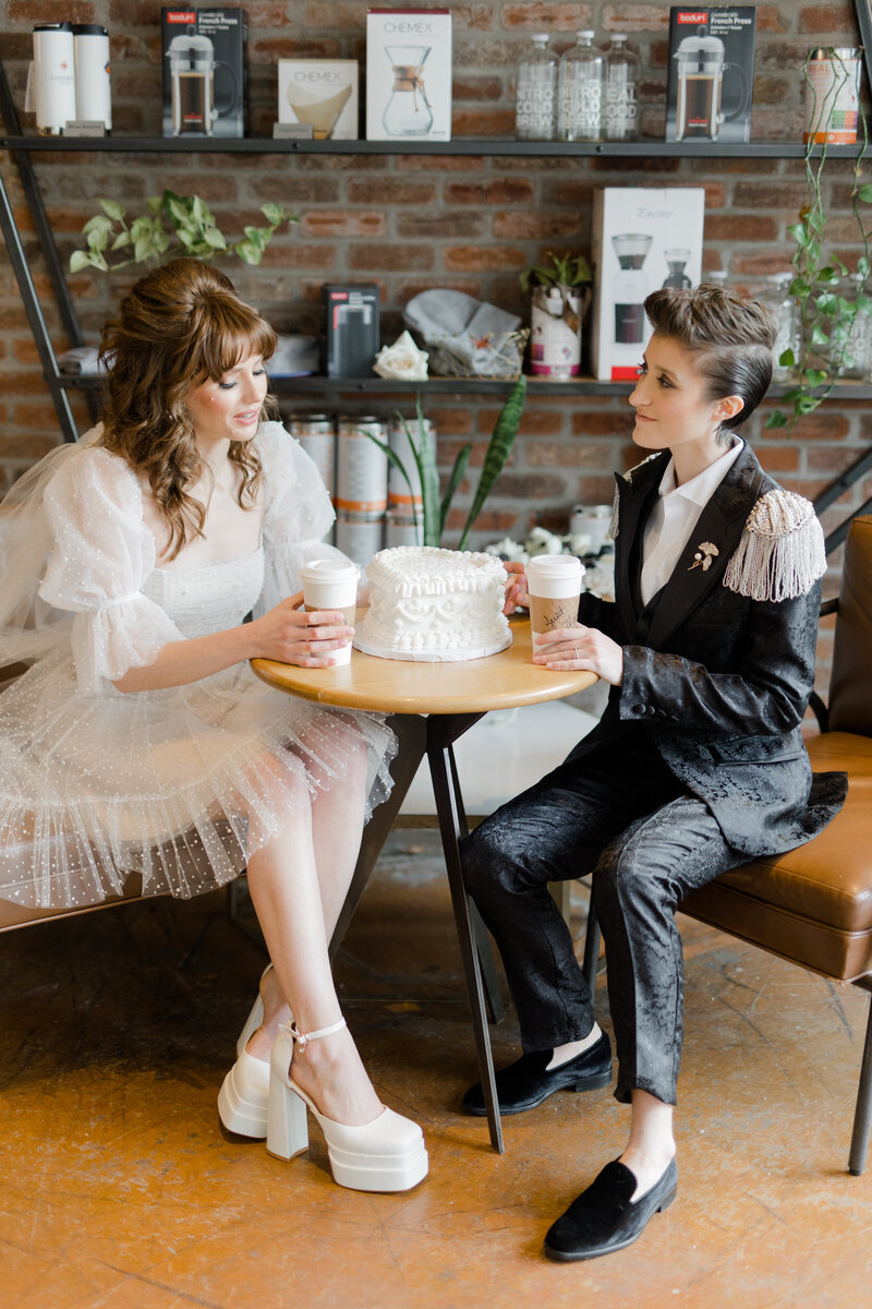Nonbinary wedding posing sitting at a table holding togo coffee cups with a white heart shaped one-tier vintage piped wedding cake resting on the table at Compass Coffee in Georgetown DC 