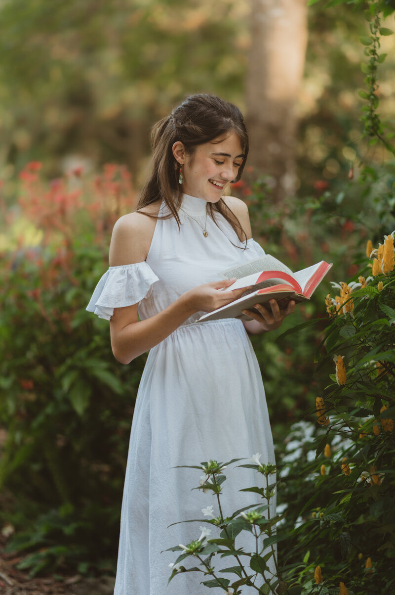 Senior girl in white dress reading a book in a garden in Winter Park, FL during her senior photo shoot