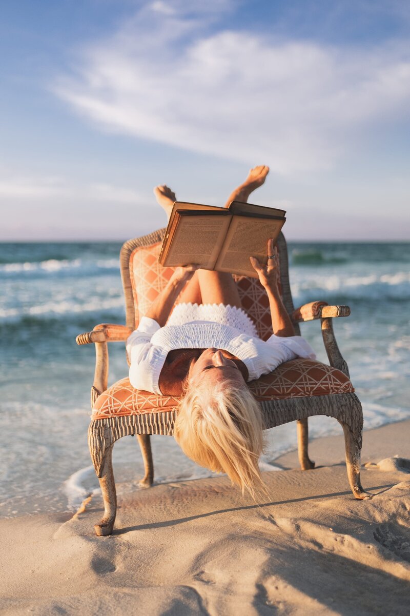 Older woman with white hair wearing a white long-sleeve dress sits upside down on an antique chair on a Florida beach at sunset while reading a book.