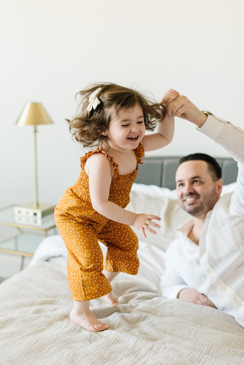 Little girl in polka dot jumpsuit bouncing on bed while holding dad's hand - Northern Virginia Family Photographer