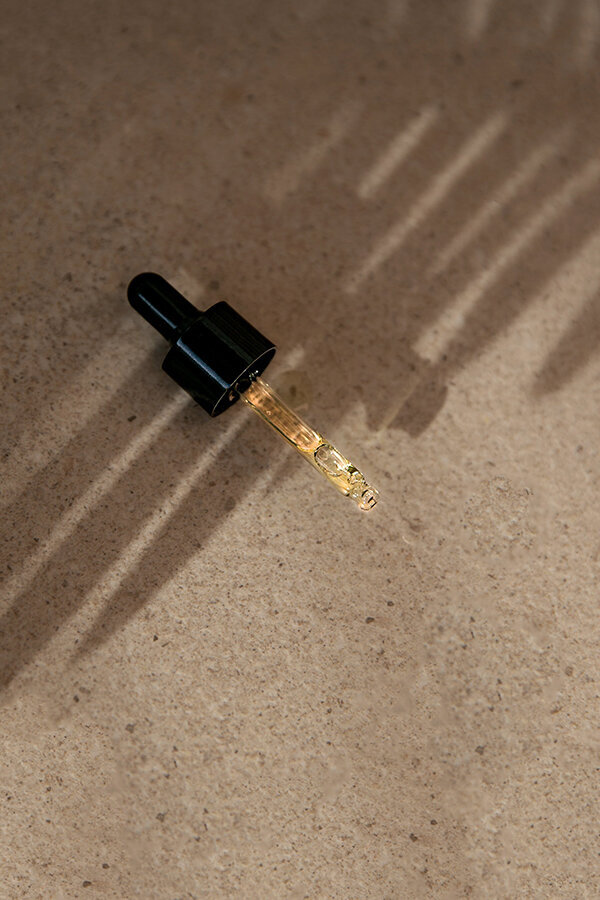 Pipette noire renfermant de l'huile essentielle doré posée sur granit à l'ombre d'un feuillage.