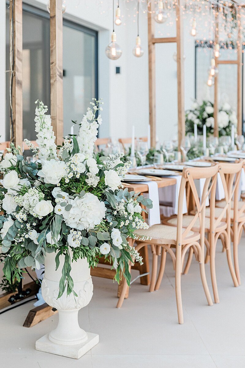Large white floral arrangement with roses, snapdragons, and eucalyptus beside a natural wood reception table set with white linens and candles