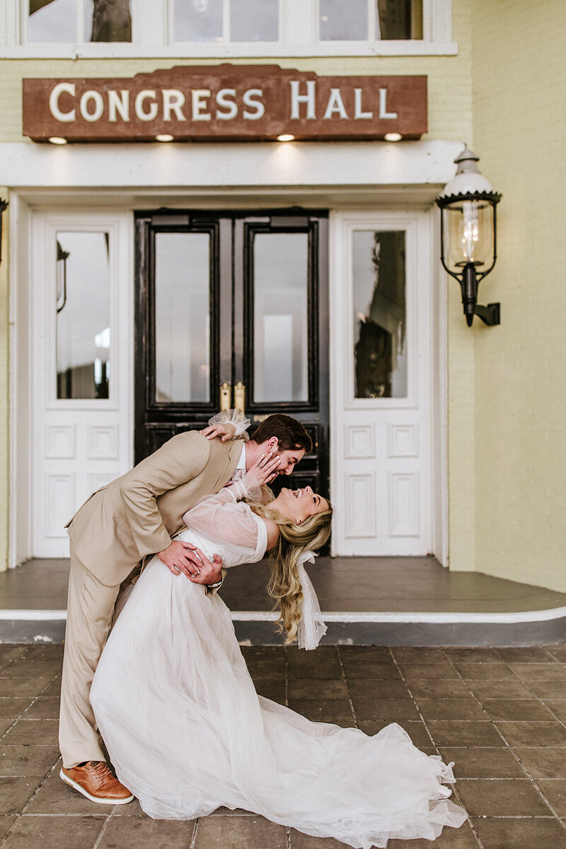 Groom in tan suit, playfully dipping bride for a kiss while she laughs with joy, in front of the Congress Hall entrance in Cape May, New Jersey.