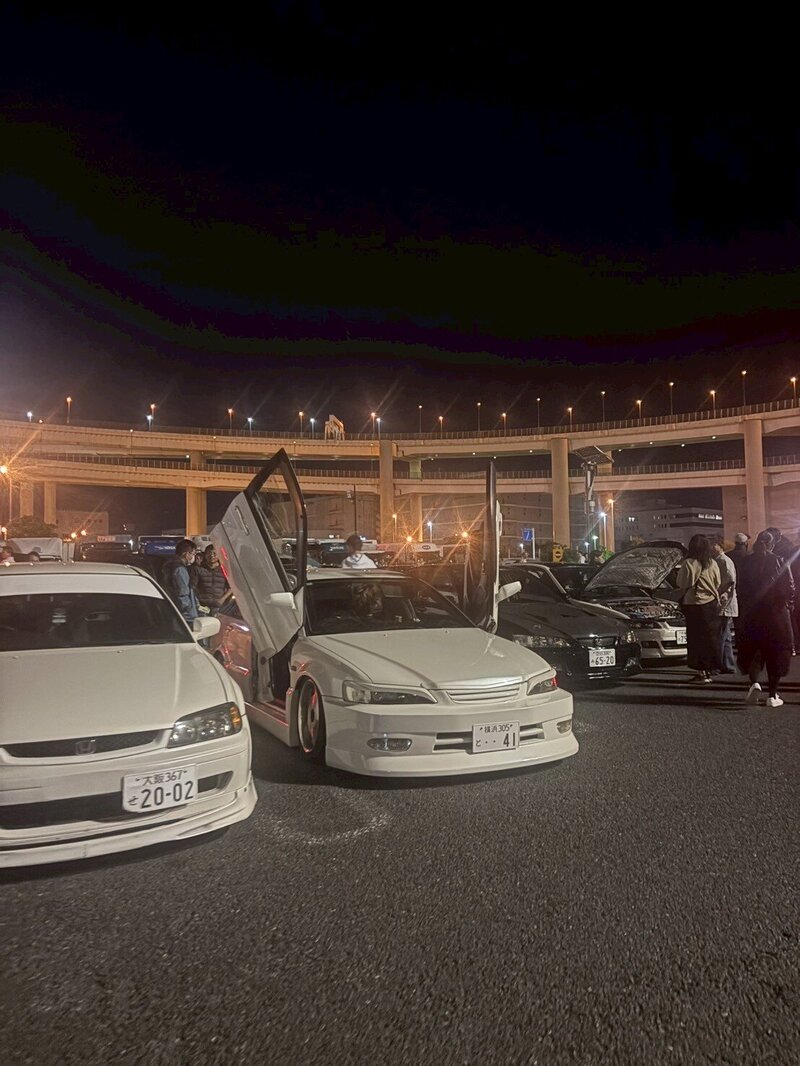 A pair of custom white cars with butterfly doors open at a nighttime car meet under the Daikoku Parking Area overpasses, surrounded by enthusiasts.
