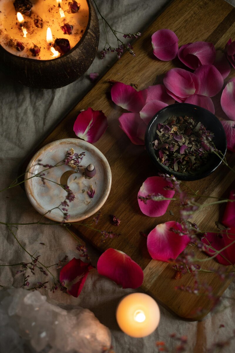 A flatlay of pink flower petals, bowls of maroon and olive green dried flowers, a large candle  and a little tea candle on top of a wood cutting board