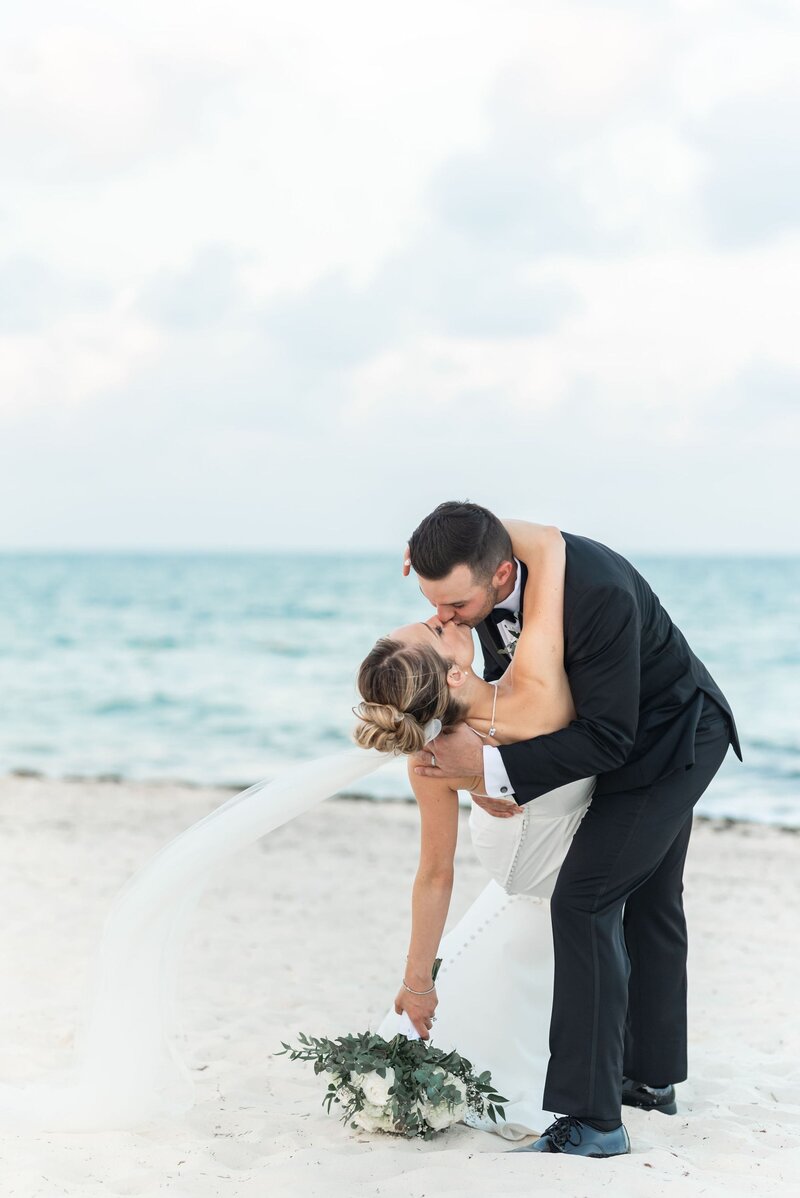 Groom dips bride into a romantic kiss along the shoreline of a tropical beach with turquoise waves and soft white sand.