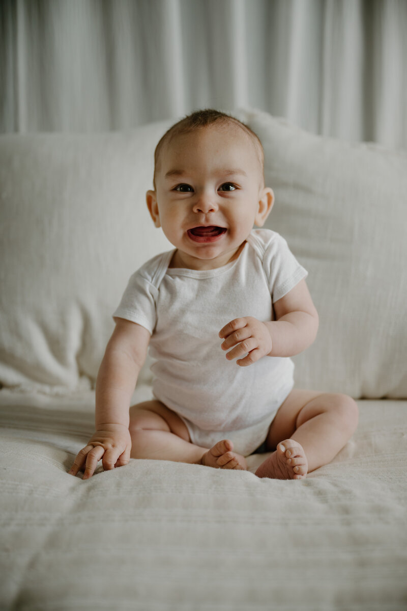 Baby boy in white smiling and looking at camera while sitting on white bed in Annapolis Maryland studio photographed by Bethany Simms Photography