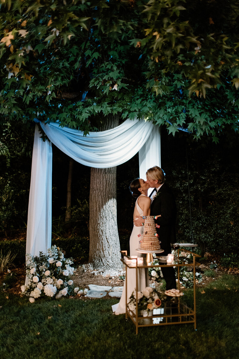 12-radiant-love-event-2021-outdoor-reception-bride-groom-kissing-cake-cutting-white-flowy-draping-tree-backdrop-romantic-elegant-timeless