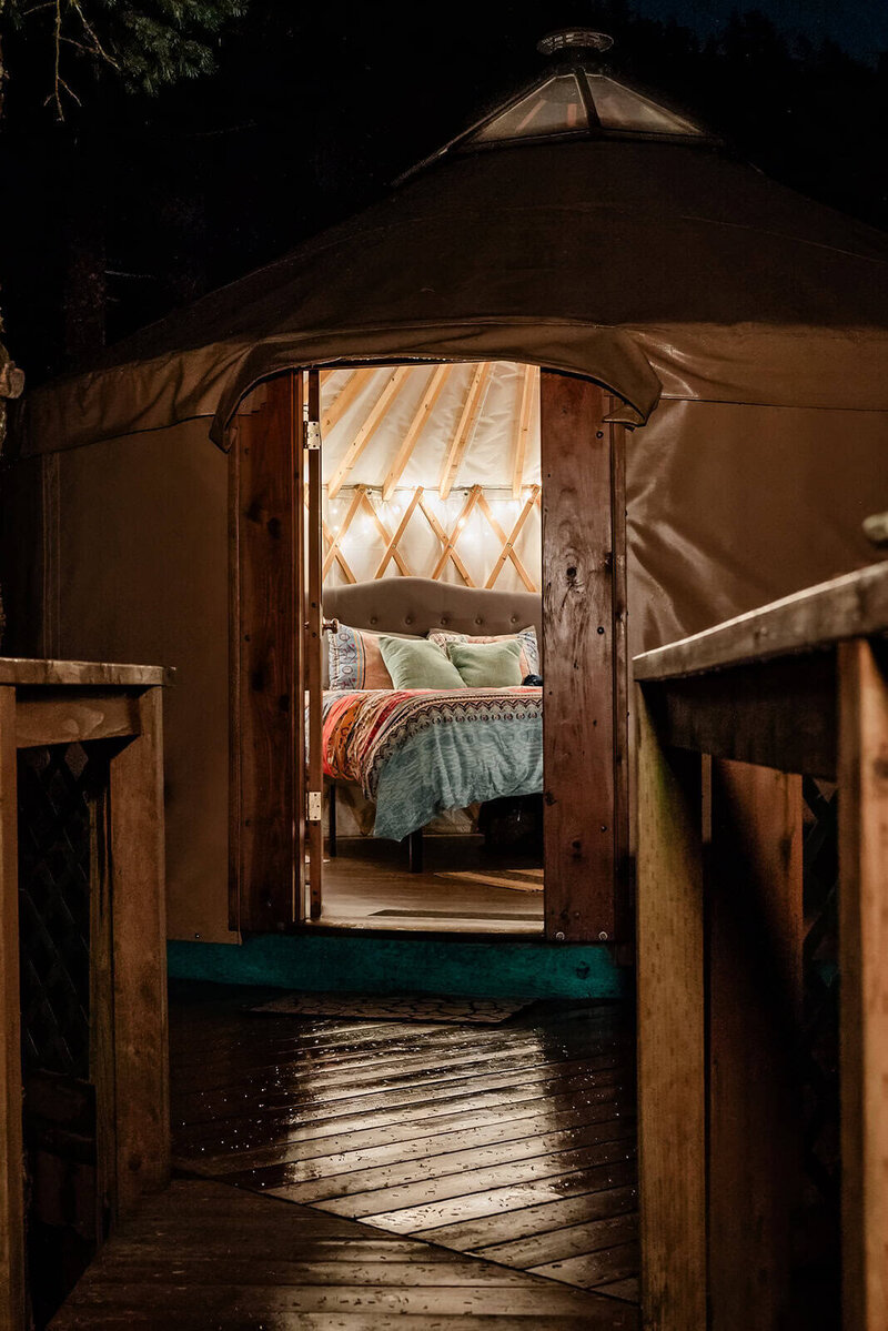 A light colored luxury canvas glamping yurt and its wodden deck are tucked into the trees at Shearwater Cove in the Kenai Fjords.    It is dark out, and the yurt is glowing with warm light out the open door as the couple inside ends their adventure elopement day.  