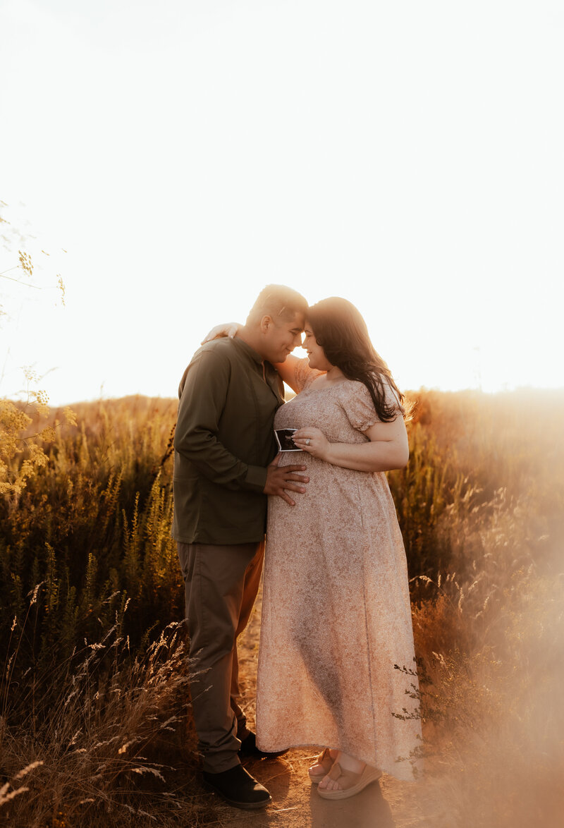 An Expecting couple stand forehead to forehead in a golden field holding their sonogram photo. 