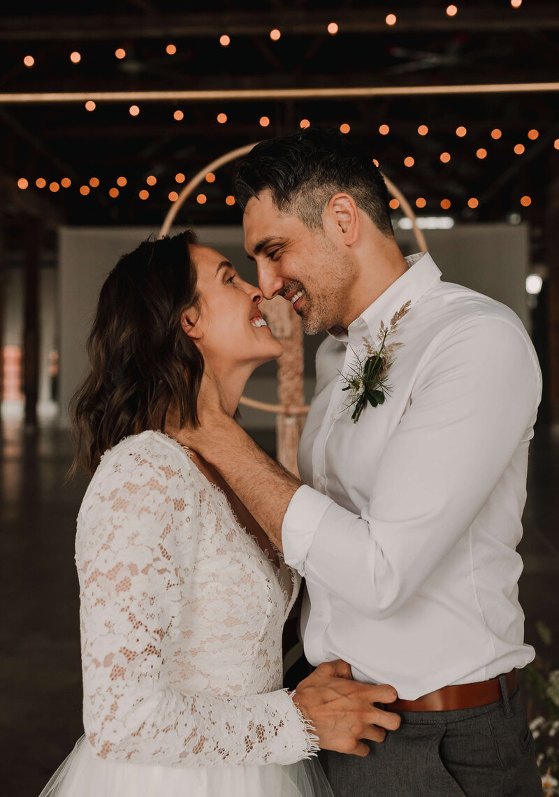 Couple smiling and holding each other at their wedding with string lights overhead.