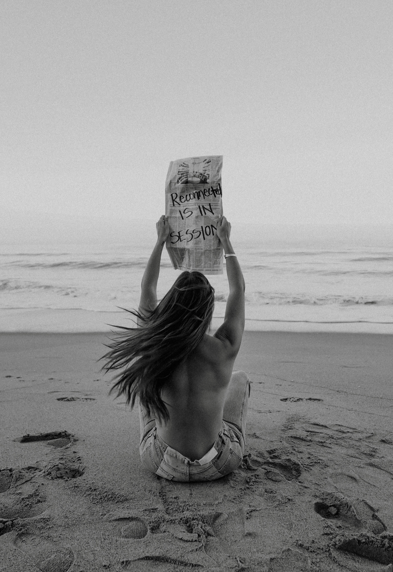 A woman sitting on the beach holding up a newspaper that says Reconnected is in session.