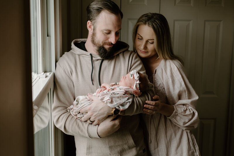 Newborn photo of dad in tan cradling baby girl in pink flower blanket with mom leaning on dad and looking over his shoulder at baby.