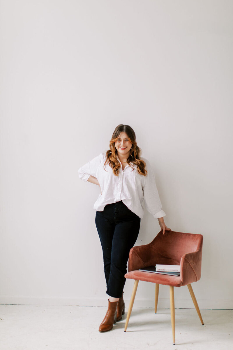 Amber is stnading beside a pink velvet chair and wearing a long sleeve white button up with black pants and brown boots