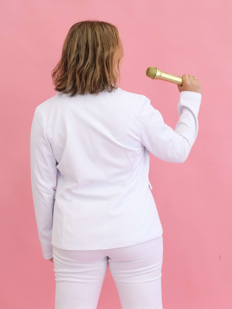 A person with shoulder-length blonde hair, dressed in a white jacket, stands facing a pink background and holds a gold microphone up with their right hand.