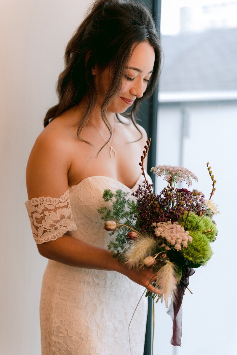 A bride in an off-shoulder lace gown holding a bouquet of wildflowers.