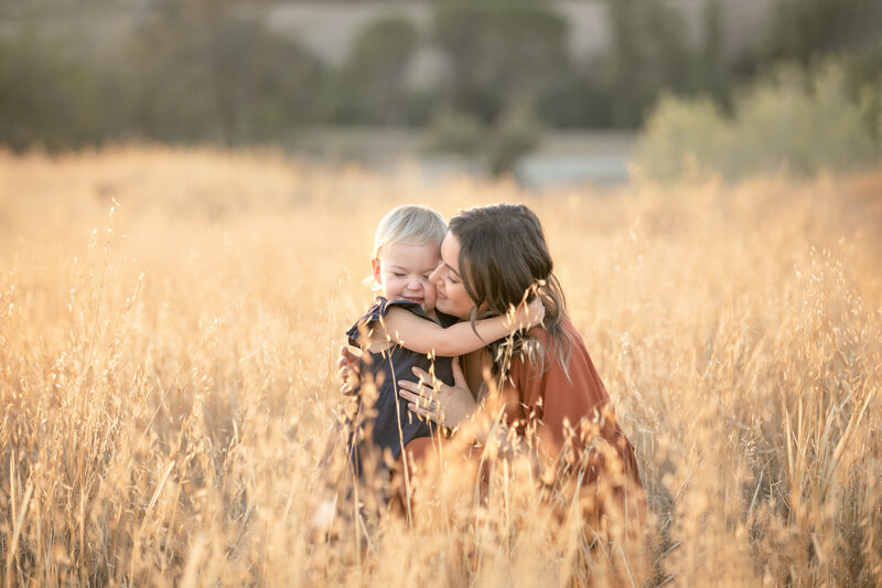 Mother daughter snuggling in a warm embrace in a field of golden grass at sunset in Fresno, CA with local family photographer