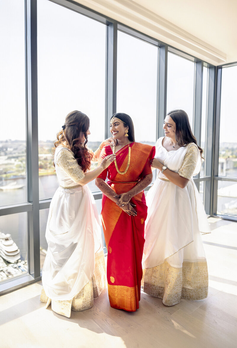 Three women stand by a large window. The central woman wears a vibrant red saree with gold jewelry, while the other two wear white and gold sarees. They are smiling and standing in a sunlit room with a cityscape visible outside.