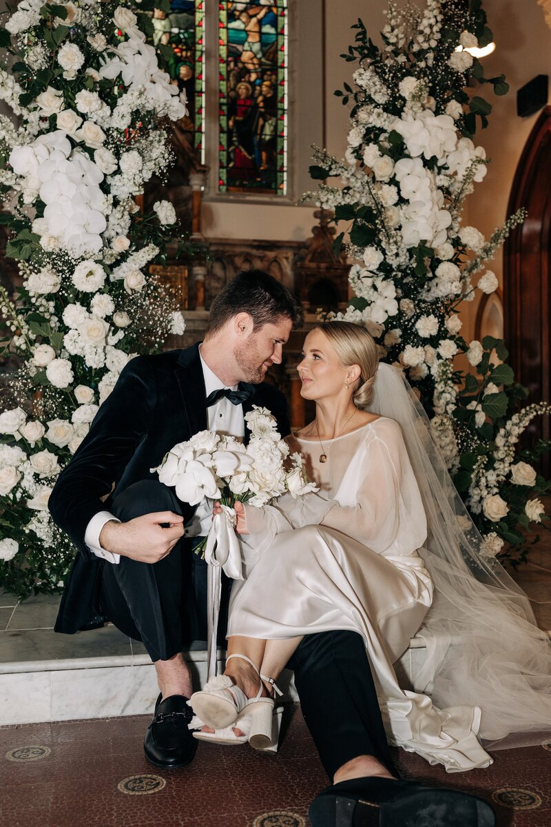 a couple in loeffler randall shoes sit in front of pillars of white flowers by the flower press at their christchurch wedding